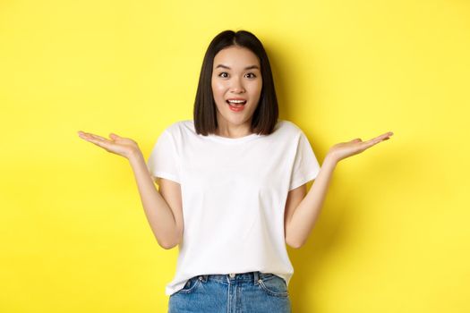 Beauty and fashion concept. Surprised asian girl spread hands sideways, holding on palms, pointing at two products, standing over yellow background.