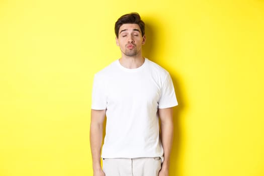 Image of lovely man close eyes and pucker lips, waiting for kiss, standing in white clothes against yellow background. Copy space