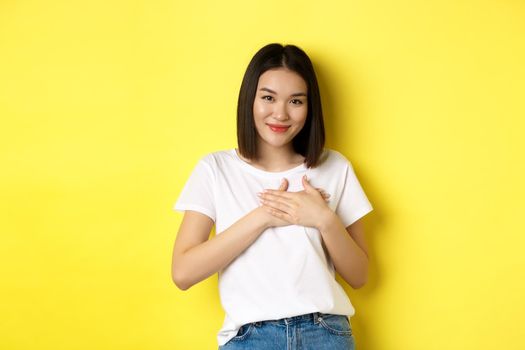 Beauty and fashion concept. Heartfelt asian girl holding hands on heart and smiling touched, thanking you, standing over yellow background.