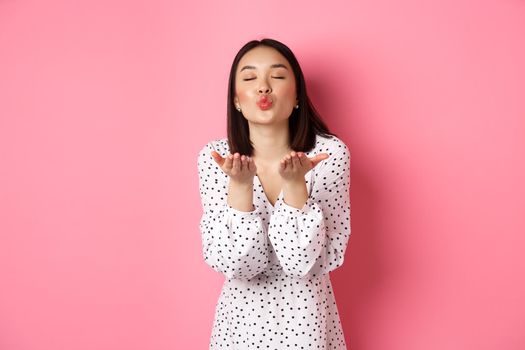 Beautiful chinese girl in dress blowing air kisses, sending mwah at camera, standing over pink background.
