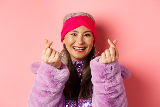 Fashion. Beautiful and fashionable asian senior woman smiling, showing korean heart gesture and looking happy, standing over pink background.