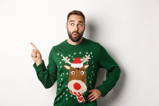 Winter holidays and christmas. Man looking curious at New Year shopping promo, standing in funny sweater against white background.