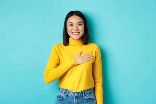Image of proud smiling asian woman holding hand on heart, showing respect to national anthem, standing over blue background. Copy space