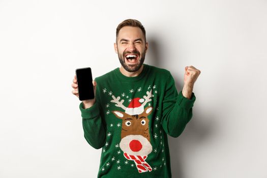 Christmas holidays and shopping concept. Excited bearded man showing mobile screen and rejoicing, winning in internet, standing over white background.