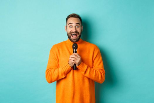 Happy adult man singing karaoke, holding microphone and looking at camera, standing in orange sweater against turquoise background.