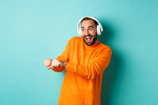 Happy man listening music in headphones and dancing funny, standing over turquoise background. Copy space