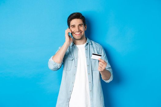 Handsome man calling bank and holding credit card, having mobile conversation, standing over blue background.