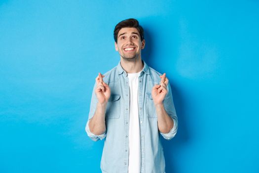 Lucky guy praying and making wish with crossed fingers, looking up with pleading face, standing against blue background.