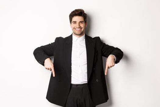 Portrait of good-looking stylish man in black suit, pointing fingers down and smiling, showing winter holidays promo, standing over white background.