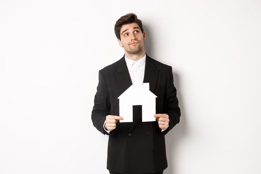 Image of handsome businessman in black suit, looking for home, holding house maket and gazing dreamy at upper right corner, standing against white background.