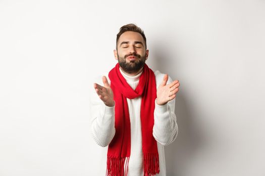 Handsome man in christmas sweater standing with closed eyes, puckered lips, leaning for kiss, standing over white background.