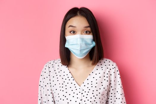 Covid-19, pandemic and lifestyle concept. Surprised asian woman in face mask raising eyebrows, staring at camera amazed, standing against pink background.