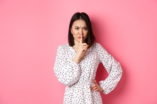 Angry asian female model shushing, frowning and hushing, showing taboo gesture, standing displeased against pink background, demand silence.