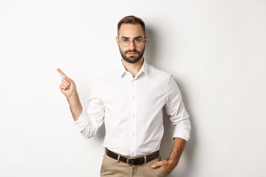 Serious bearded man pointing finger left, showing advertisement, standing over white background.