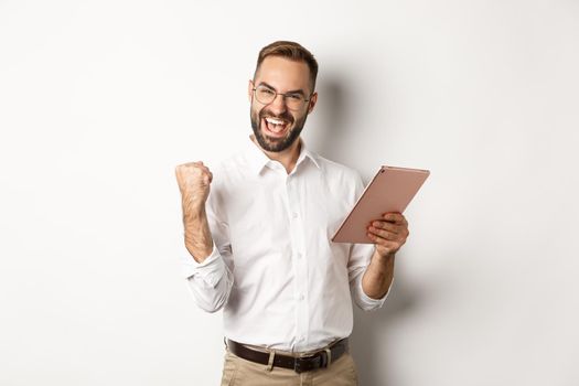 Successful businessman rejoicing on winning online, reading on digital tablet and making fist pump, triumphing, standing over white background.