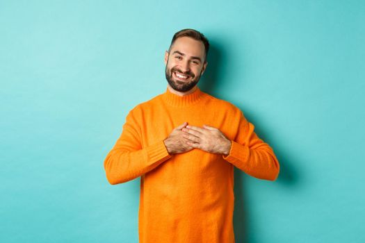 Grateful man smiling, holding hands on heart, thank you gesture, feeling touched with gift, standing over turquoise background.