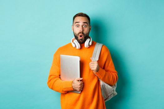 Happy man with backpack and headphones, holding laptop and smiling, looking surprised, standing over turquoise background.