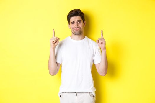 Unamused handsome guy frowning, pointing fingers up at something bad, standing skeptical against yellow background.