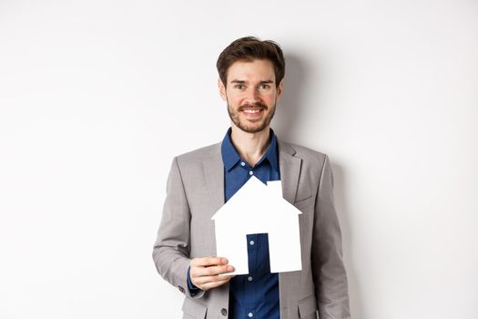 Real estate. Handsome young businessman buying property, holding paper house cutout and smiling, agency advertisement, white background.