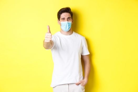 Concept of coronavirus, pandemic and social distancing. Confident young man in medical mask showing thumbs up and winking, yellow background.