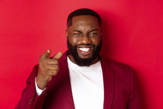 Cheerful Black man with beard wearing party clothes, pointing finger at camera and winking, congratulate with christmas and new year, standing over red background.