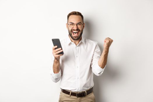 Successful businessman looking happy, fist pump and rejoice in winning online lottery, standing over white background.