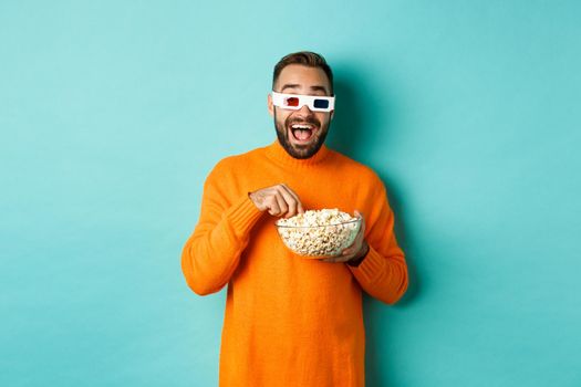 Happy and amazed young man in 3d glasses watching comedy movie, looking at tv screen and eating popcorn, standing over blue background.