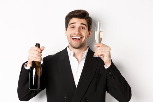 Close-up of handsome man in suit, holding bottle and glass of champagne, celebrating holiday, standing against white background.
