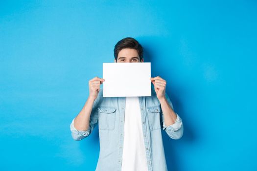 Sneeky handsome guy hiding face behind blank piece of paper for your logo, making announcement or showing promo offer, standing over blue background.