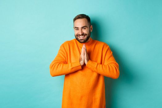 Happy man saying thank you, holding hands in pray, looking grateful and smiling, standing over light blue background.