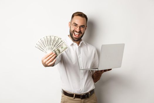 Business and e-commerce. Happy successful businessman bragging with money, working on laptop online, standing over white background.