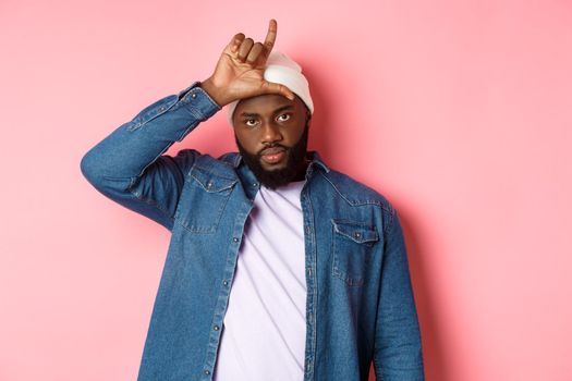 Sad african-american man showing loser sign on forehead and staring at camera, standing over pink background.