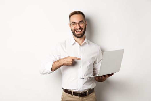 Business. Handsome manager in glasses working on laptop, pointing at computer and smiling pleased, standing over white background.