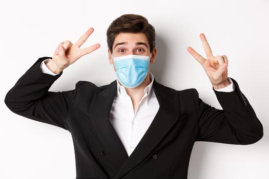 Concept of covid-19, business and social distancing. Close-up of happy businessman in suit and medical mask, showing peace signs and smiling, standing against white background.