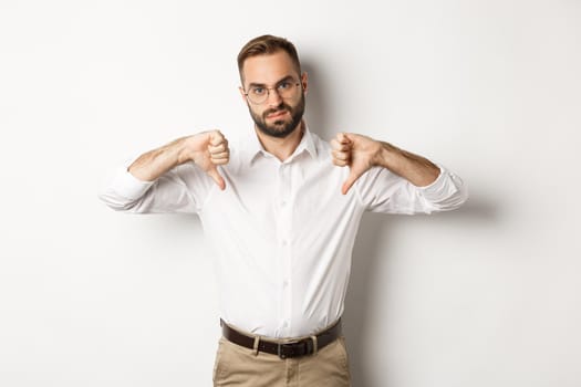 Displeased businessman in glasses showing thumbs down, dislike and disapprove, standing over white background.