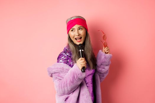 Gorgeous asian senior woman singing karaoke in mic, perform song and looking happy, standing over pink background.