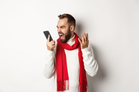 Angry man shouting at smartphone with mad face, standing furious against white background.