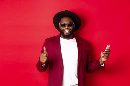 Handsome african american man using phone app and showing thumb up, smiling at camera, wearing sunglasses and fancy hat, red background.