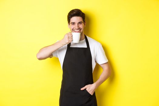 Cafe barista drinking cup of coffee and smiling, wearing black apron, standing over yellow background.