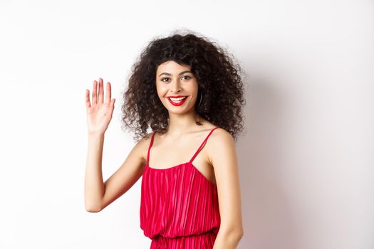 Beauty and fashion. Friendly elegant woman with curly hair, red dress, waving hand and saying hello, smiling to greet you, standing on white background.