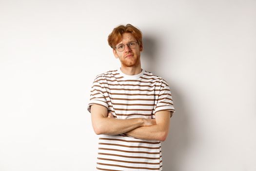 Teenage redhead guy in glasses cross arms on chest, looking skeptical and unamused at camera, standing over white background.