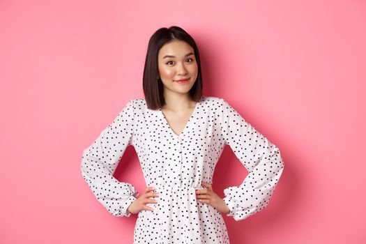 Beautiful korean lady in dress looking at camera and smiling, waiting for something, standing against pink background.