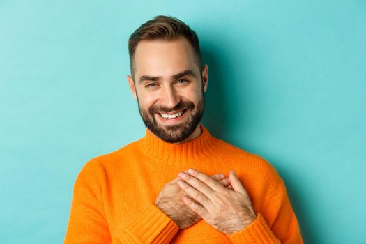 Close-up of handsome young man saying thank you, holding hands on heart and smiling grateful, standing over light blue background.