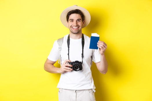 Travelling, vacation and tourism concept. Smiling man tourist holding camera, showing passport with tickets, standing over yellow background.