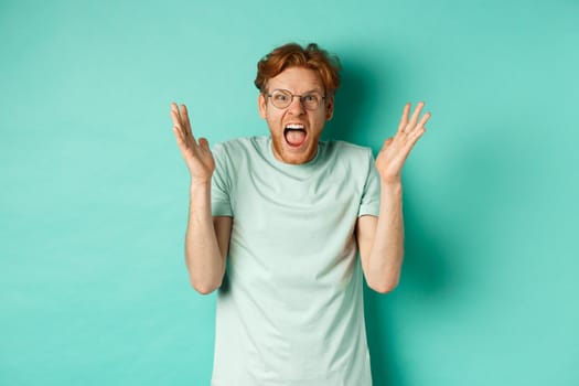 Portrait of distressed and angry redhead guy losing temper, shouting and shaking hands outraged, staring with furious face at camera, standing over mint background.