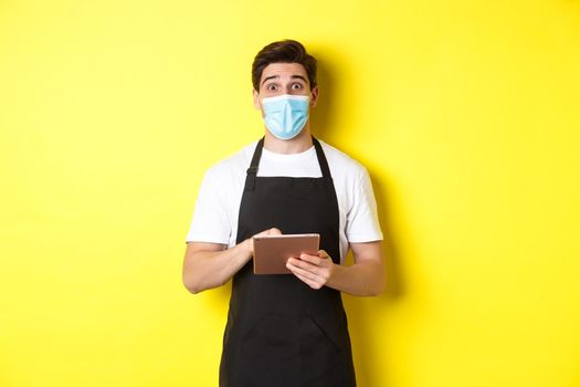 Concept of covid-19, small business and pandemic. Waiter in black apron and medical mask taking order, holding digital tablet, standing over yellow background.