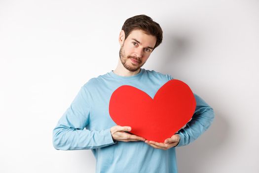 Romantic boyfriend making Valentines day surprise, holding big red heart cutout on chest and smiling with love, looking tender at camera, standing over white background.