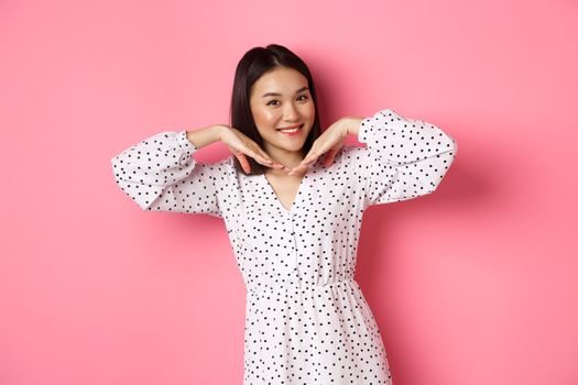 Lovely asian woman showing her beautiful clean face, smiling pretty, standing in dress against pink romantic background.