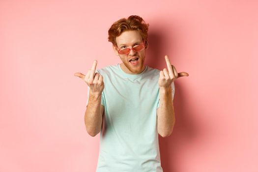 Arrogant and careless redhead man in sunglasses dont give a fuck, showing middle fingers at camera and frowning, standing over pink background.