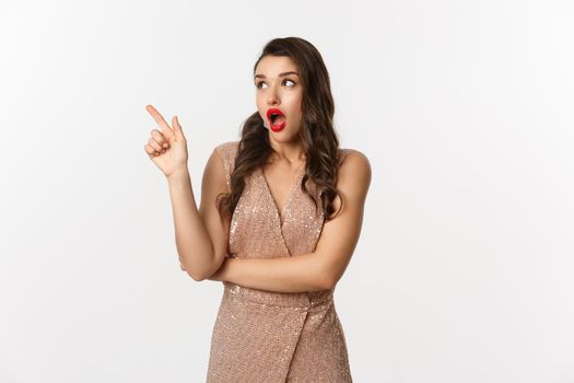 Christmas, holidays and celebration concept. Amazed fashionable woman in party dress and red lipstick, pointing and looking left at promo offer, standing against white background.